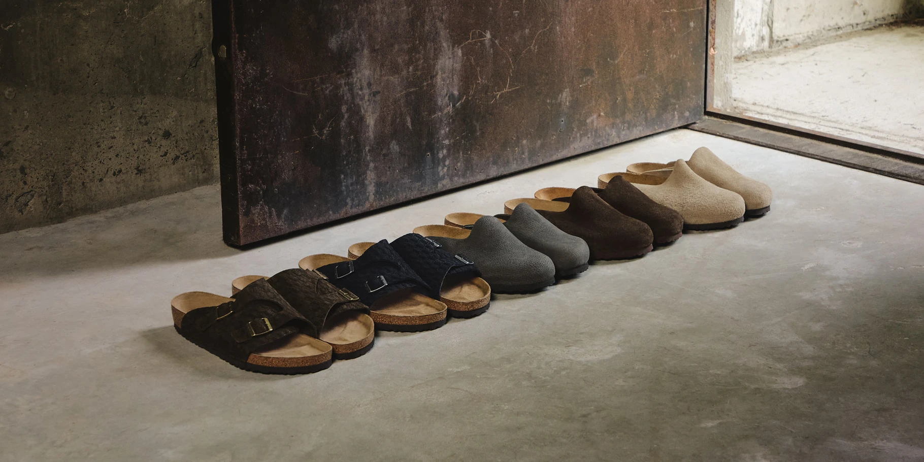 The Kith for Birkenstock Summer 26 capsule lined up on a concrete floor: buckled Zurich sandals in front, closed-toe Amsterdam clogs in Mocca, Taupe, and Black behind them